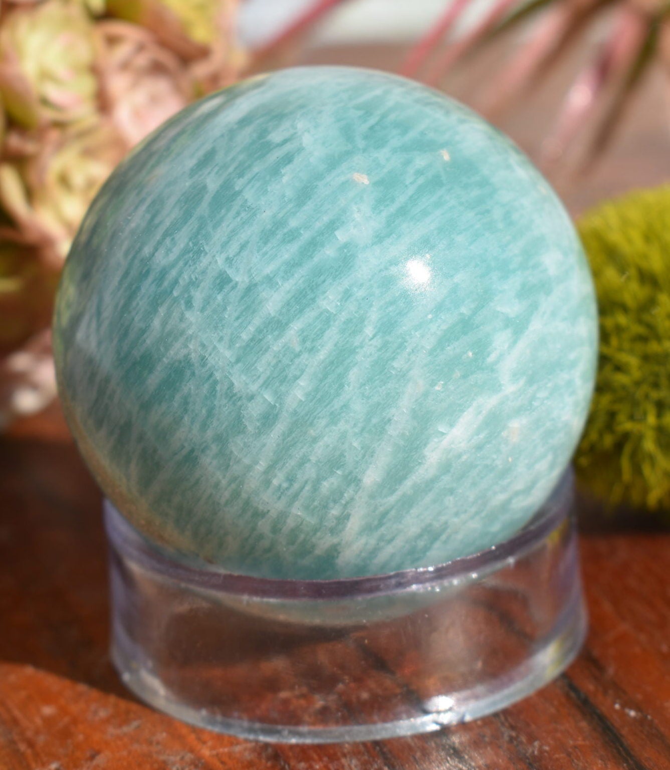 A light green sphere of amazonite sits perched atop a clear glass ring, on a wooden surface. The background of the image contains a succulent bouquet and ball of moss.