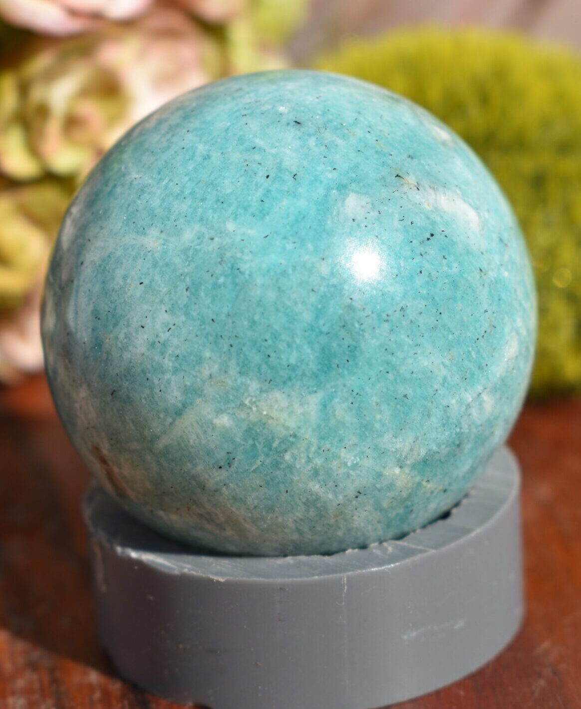 A sphere of light green amazonite sits on top of a grey ring, on a wooden surface. There is a moss ball and a succulent bouquet in the background.