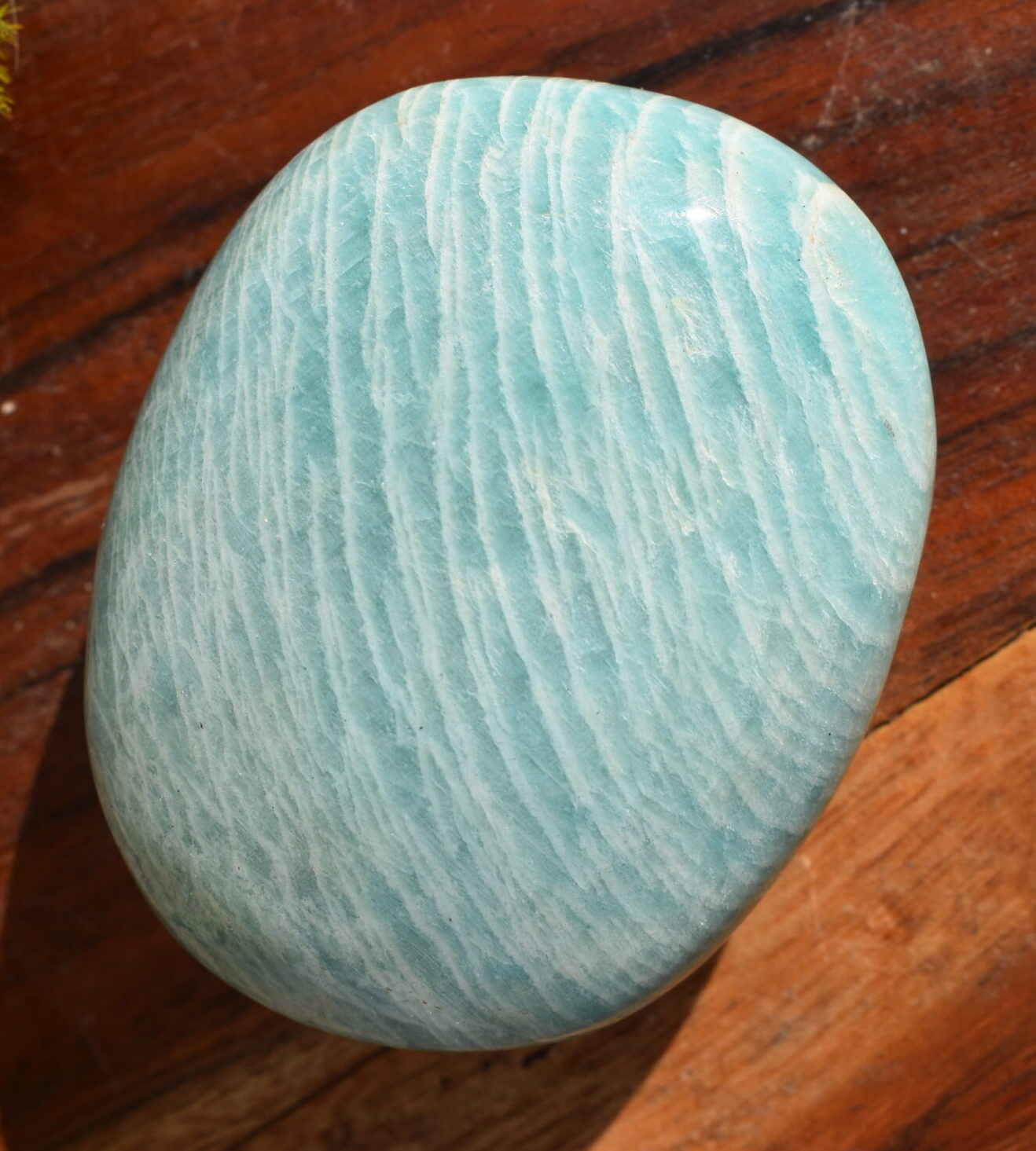 A piece of light green amazonite, in the shape of a squared off oval, sits atop a wooden surface.