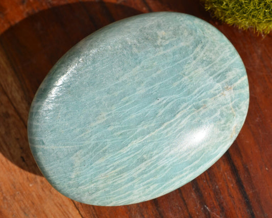 A rounded oval shaped piece of light green amazonite sits atop a wooden surface. There is a small piece of moss in the top right hand corner of the image.